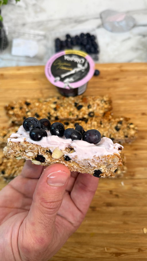 Blueberry granola bars topped with @yoproau limited edition Smoothie Bowl flavour - the ultimate grab-and-go snack. Hurry, this flavour is only here for a very limited time!
Ingredients: 
For the granola bars: 
- 2 cups rolled oats 
- 1/2 cup shredded coconut 
- 1/2 cup dried or freeze-dried blueberries 
- 1/3 cup raw almonds, roughly chopped 
- 1/4 cup sunflower seeds and pumpkin seeds
- 1/4 teaspoon salt 
- 1 teaspoon ground cinnamon 
- 1/3 cup natural peanut butter 
- 1/3 cup honey + 1–2 extra tablespoons if mixture looks dry 
- 1 tablespoon neutral oil (coconut, light olive, canola, or a knob of melted butter) 
- 1 teaspoon vanilla extract For the yoghurt topping: 
- 1/2 cup YoPRO limited edition Smoothie Bowl flavour yoghurt 
- 1/4 cup fresh blueberries 
1. Preheat the oven to 160°C.
2. Line a 20x20cm baking pan with parchment paper.
3. In a large bowl, combine oats, shredded coconut, dried blueberries, nuts, seeds, salt and cinnamon.
4. In a small saucepan over low heat, gently warm the peanut butter, honey, and oil until smooth and runny. Remove from heat and stir in vanilla extract.
5. Pour the warm mixture over the dry ingredients. Mix well to coat everything evenly. If it still looks dry, drizzle in an extra tablespoon of honey.
6. Transfer the mixture to the lined baking pan. Press it down firmly.
7. Bake for 20–25 minutes, until the edges are golden brown. Keep an eye so it doesn’t burn.
8.Cool completely in the pan on a wire rack.
9. Cut cooled granola into bars, spread with YoPRO yoghurt and blueberries.
10. Chill in the fridge for 30 minutes to help the topping set. Store in an airtight container in the fridge for up to 5 days.
#AD #FeedYourProgress #GranolaBars #GranolaRecipe