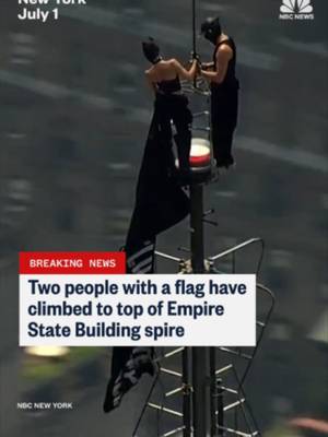 At least two people are seen on top of the Empire State Building's spire with a flag.
