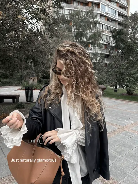 A white ruffled blouse layered under an oversized black leather jacket, paired with dark straight-leg jeans and beige suede cowboy boots. A structured brown leather shoulder bag completes the look. Voluminous soft curls and dark sunglasses add a polished, effortlessly chic touch. Serena van der Woodsen vibe, elevated casual, western-inspired, feminine, effortless, Gossip Girl aesthetic.