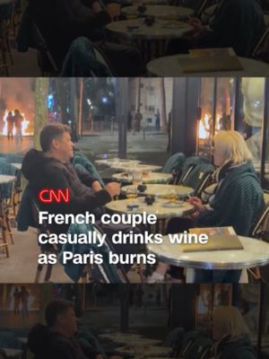 A couple were seen smoking and drinking wine near the Arc de Triomphe in Paris on Saturday night as protests unfolded in the background. Flames from a burning trash can could be seen in the distance as the two talked. #france #protests